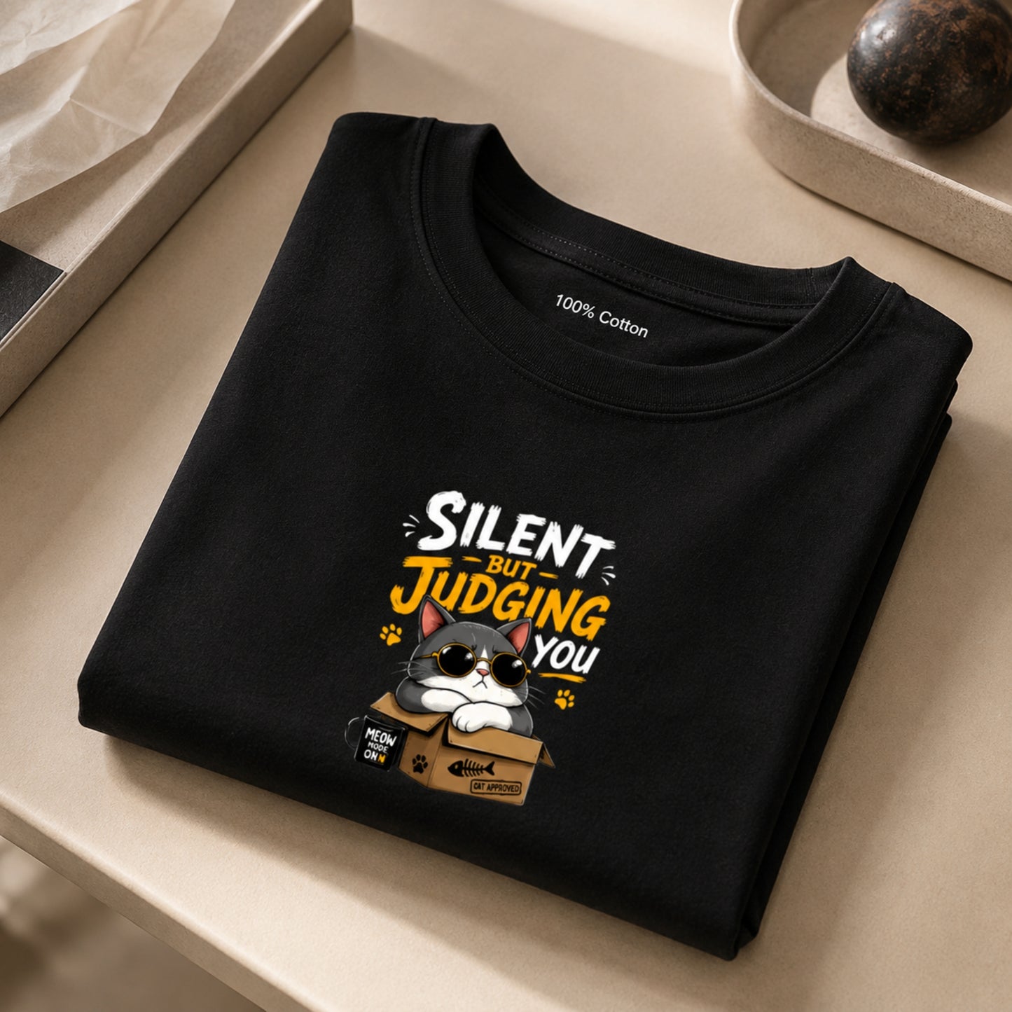 Black unisex stretch t-shirt featuring a cute sunglasses-wearing cat sitting on a box with the text “Silent But Judging You” in bold yellow and white typography, humorous meme-style graphic design on chest.