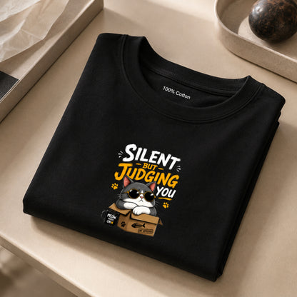 Black unisex stretch t-shirt featuring a cute sunglasses-wearing cat sitting on a box with the text “Silent But Judging You” in bold yellow and white typography, humorous meme-style graphic design on chest.