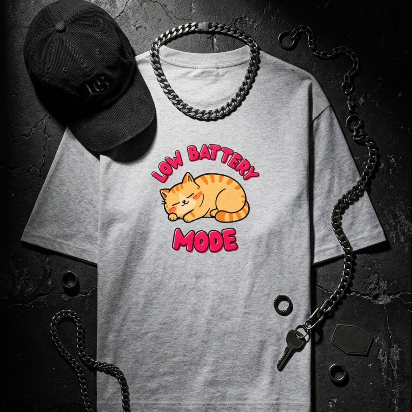 Gray t-shirt with a cat graphic and 'Low Battery Mode' text, accompanied by a black cap and chain on a dark background.
