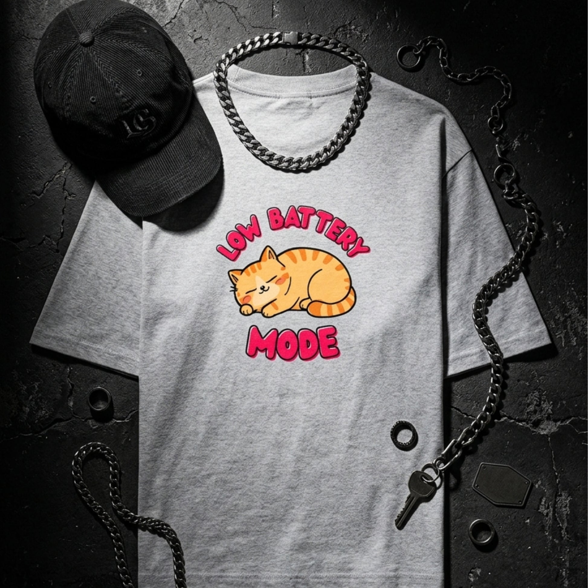 Gray t-shirt with a cat graphic and 'Low Battery Mode' text, accompanied by a black cap and chain on a dark background.