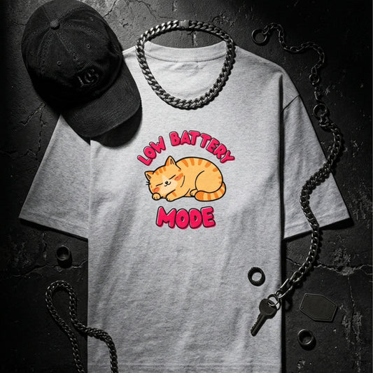Gray t-shirt with a cat graphic and 'Low Battery Mode' text, accompanied by a black cap and chain on a dark background.