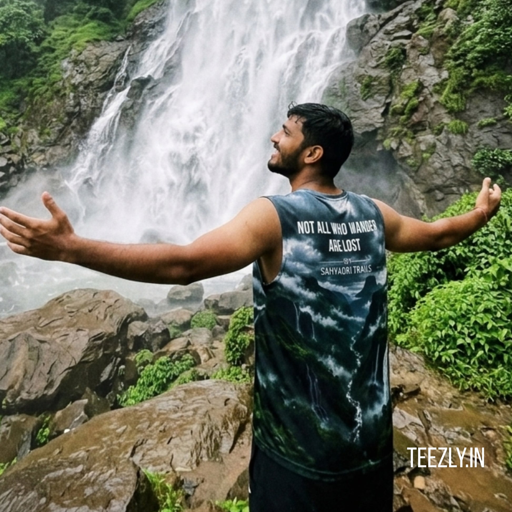 Man with arms outstretched in front of a waterfall, wearing a tank top with text.
