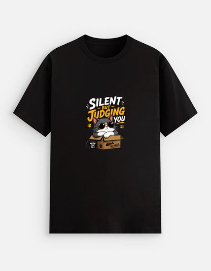 Black unisex stretch t-shirt featuring a cute sunglasses-wearing cat sitting on a box with the text “Silent But Judging You” in bold yellow and white typography, humorous meme-style graphic design on chest.